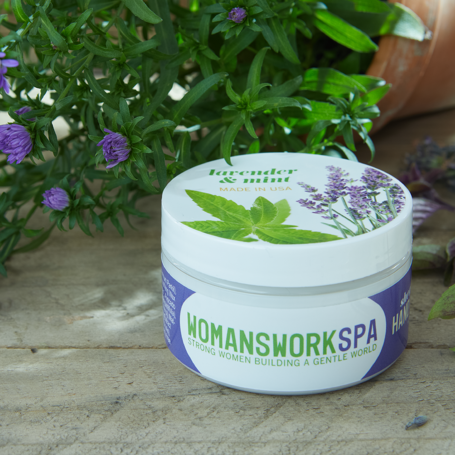Jar of 'Womanswork Spa' product with lavender and mint design on a wooden surface with plants in the background.