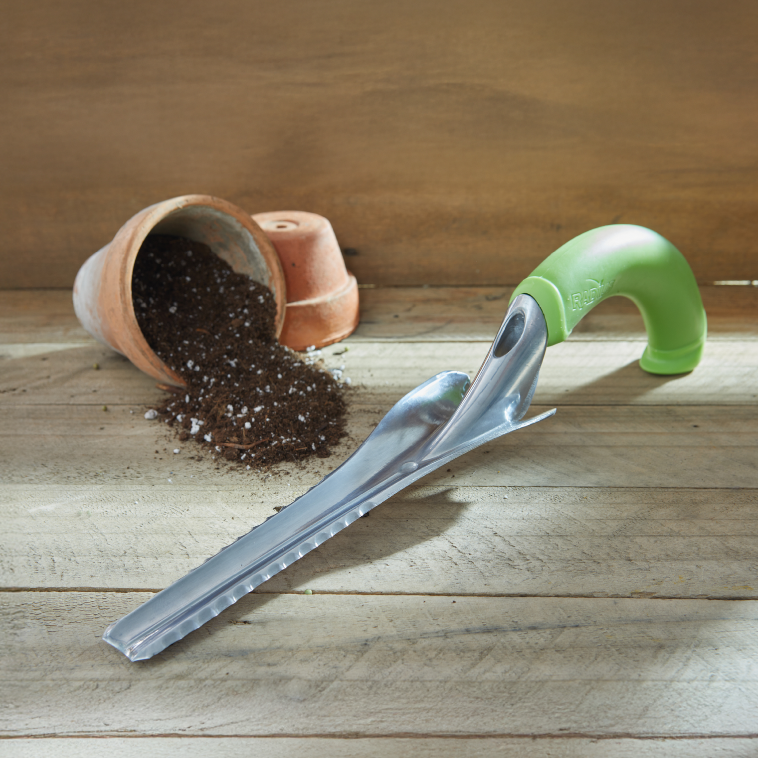 Garden tool with green handle on a wooden surface next to an overturned terracotta pot with soil.
