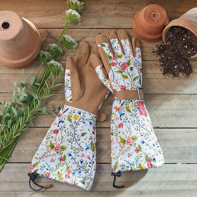 Floral gardening gloves on a wooden surface with plants and pots.