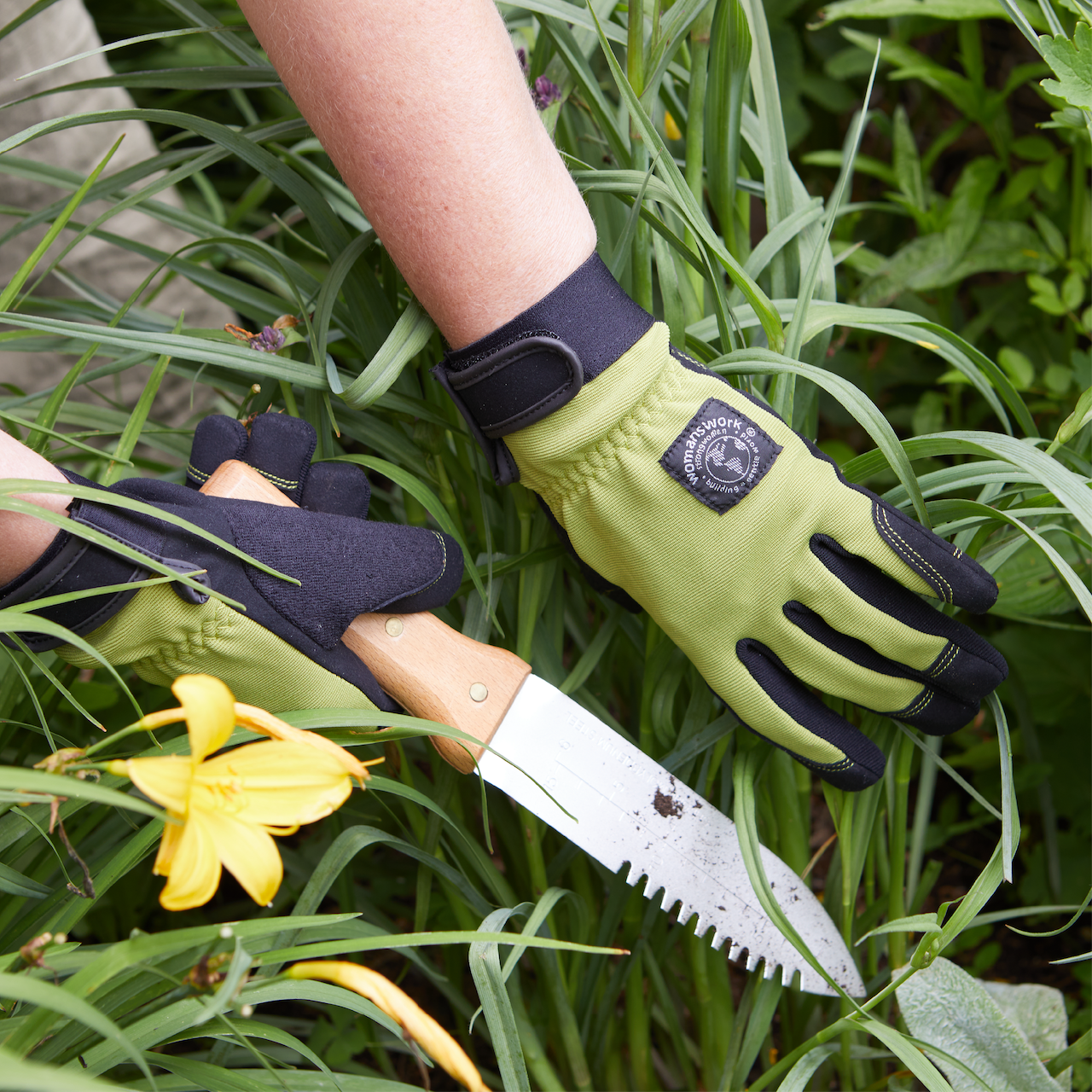 Person wearing gardening gloves holding a knife among green plants with a yellow flower.