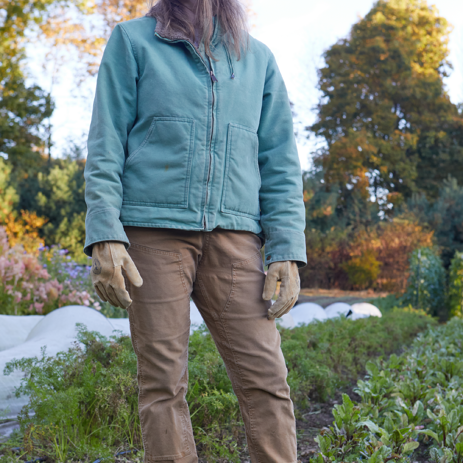 Person wearing a teal jacket and brown gloves standing in a garden.
