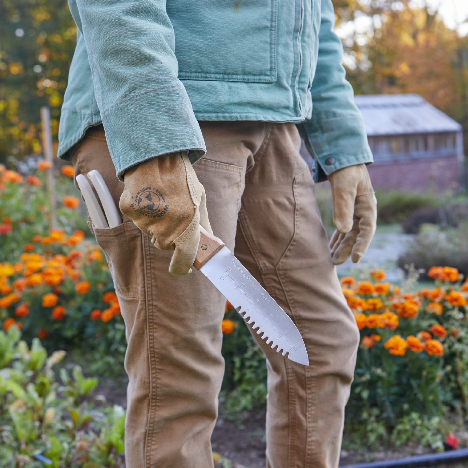 Person holding a garden tool in a garden setting with flowers and a building in the background