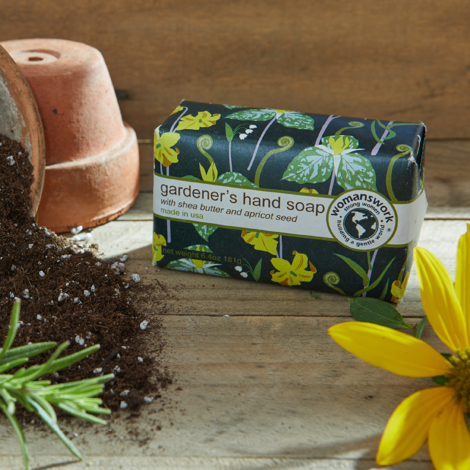 Bar of gardener's hand soap with floral packaging on a wooden surface with soil and a flower.