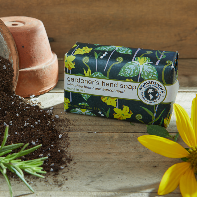 Bar of gardener's hand soap with floral packaging on a wooden surface with soil and a flower.