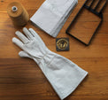 White leather glove on a wooden surface with a spool of thread, paper, and metal tool.