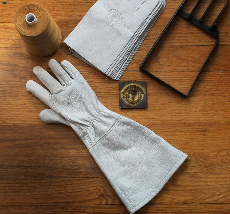 White leather glove on a wooden surface with a spool of thread, paper, and metal tool.