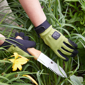 Person wearing gardening gloves holding a knife among green plants with a yellow flower.